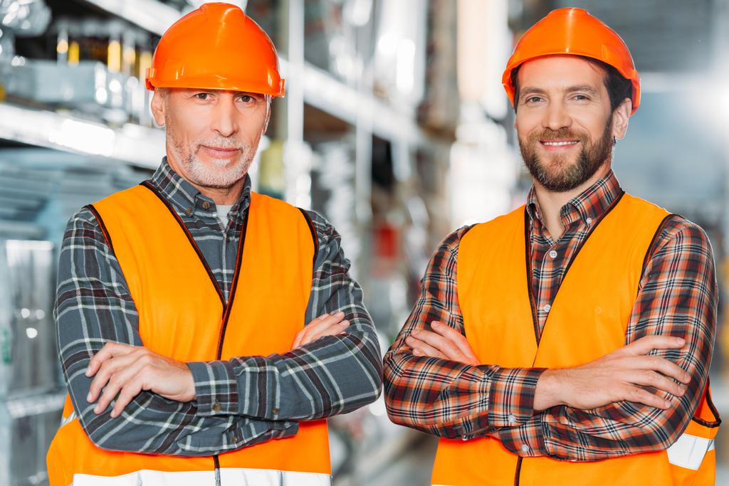 two male workers in safety vests and helmets with crossed arms in storehouse - Photo, Image