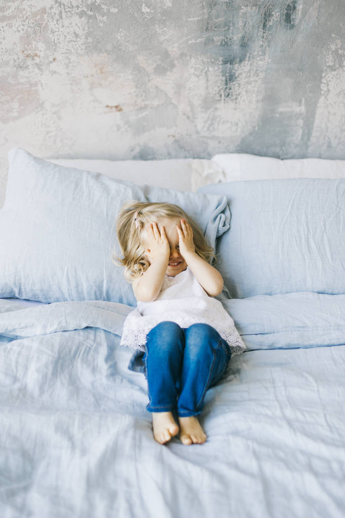 όμορφο ξανθό κοριτσάκι έκλεισε τα μάτια με τα χέρια στο κρεβάτι στο σπίτι - Φωτογραφία, εικόνα