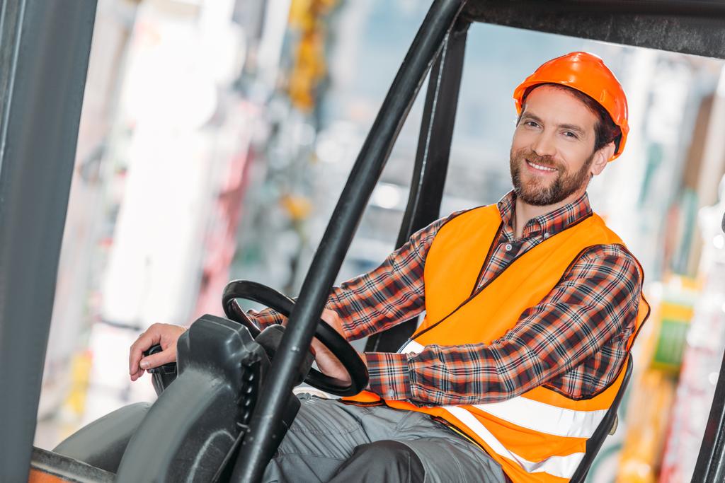 trabalhador masculino em colete de segurança e capacete sentado na máquina de empilhadeira em armazenamento
 - Foto, Imagem