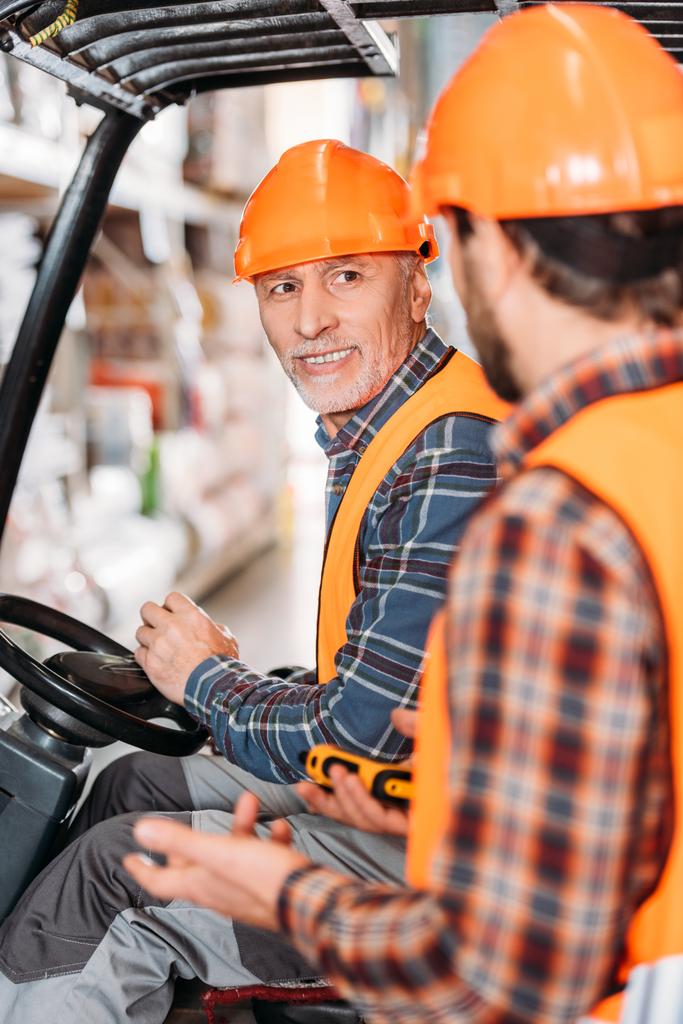 lavoratore anziano in giubbotto di sicurezza e casco seduto in macchina elevatore e parlando con il collega in magazzino
 - Foto, immagini