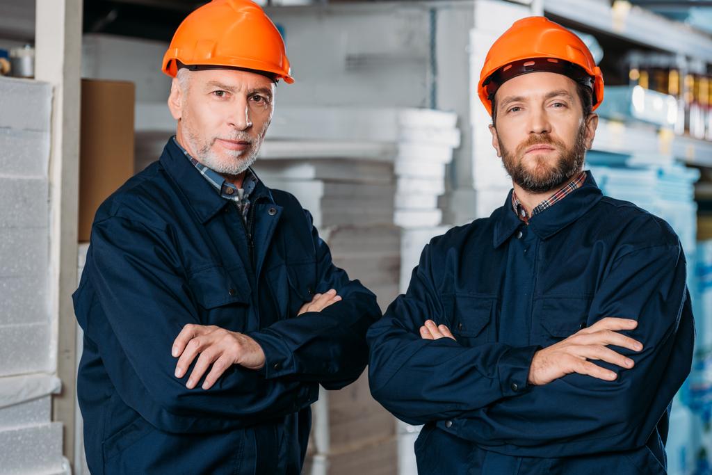 male workers in helmets posing with crossed arms in storehouse - Photo, Image
