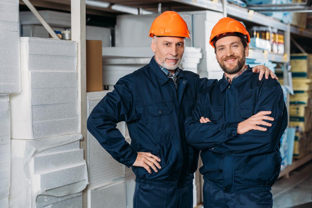 trabalhadores do sexo masculino em capacetes abraçando e posando no armazém
 - Foto, Imagem