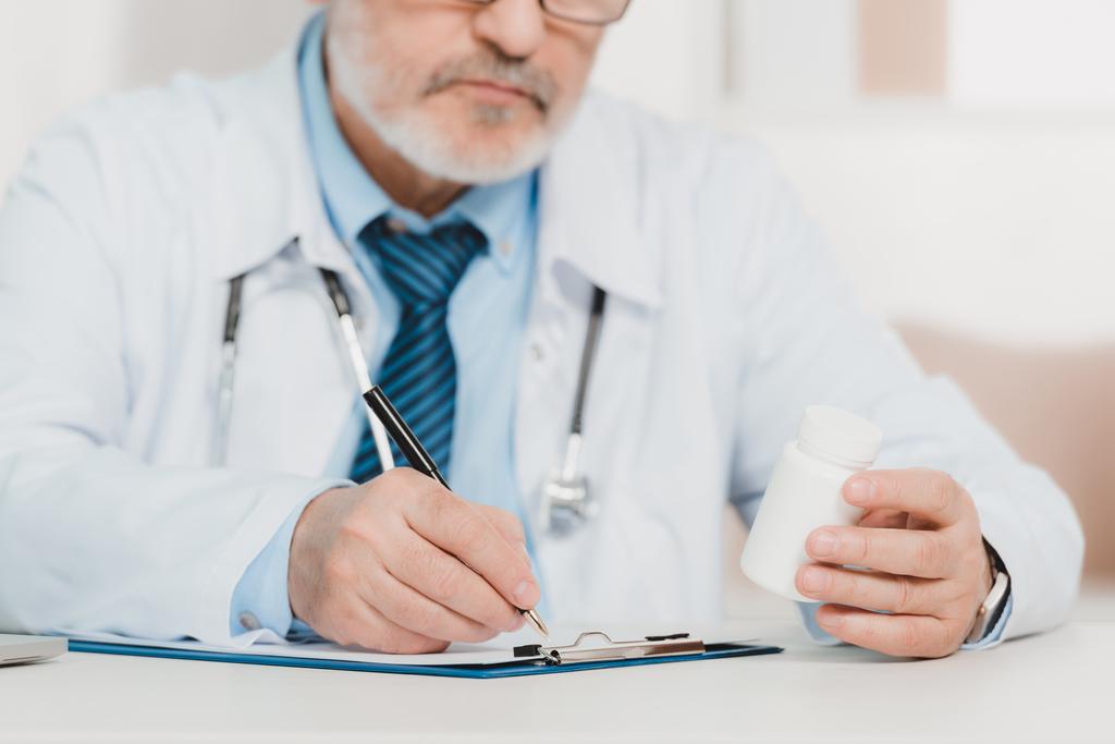 Μερική άποψη του γιατρού με χάπια στο χέρι κάνοντας σημειώσεις στο Σημειωματάριο (Notepad) στο χώρο εργασίας - Φωτογραφία, εικόνα