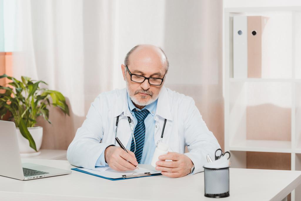 πορτρέτο του επικεντρώθηκε ανώτερος γιατρός κάνοντας σημειώσεις στο Σημειωματάριο (Notepad) στο χώρο εργασίας με το laptop στην κλινική - Φωτογραφία, εικόνα