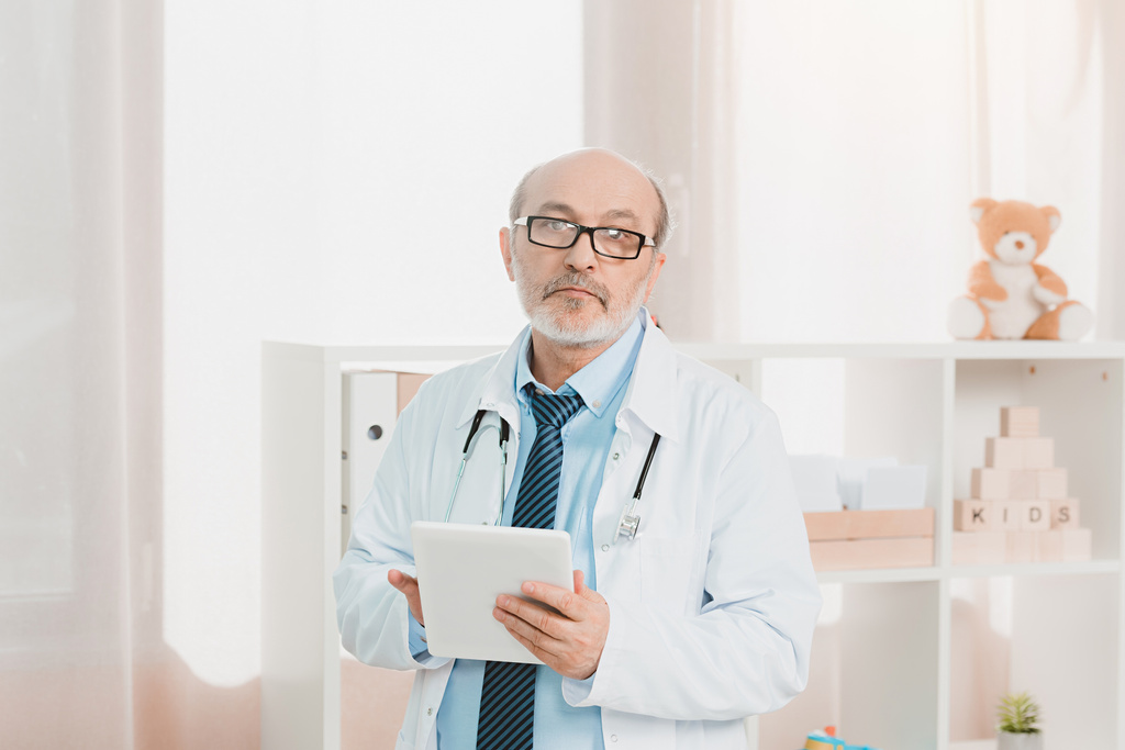 portrait of senior doctor in white coat with tablet in hands looking at camera in clinic - Photo, Image