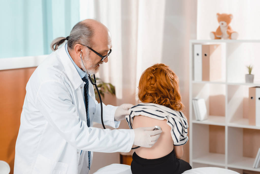 старший врач в белом халате и медицинских перчатках осматривает девушку со стетоскопом в клинике
 - Фото, изображение