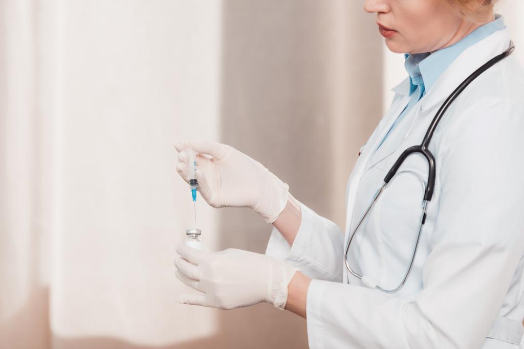 částečný pohled doktor v bílém plášti a lékařské rukavice Držte injekční stříkačky v rukou v klinice - Fotografie, Obrázek