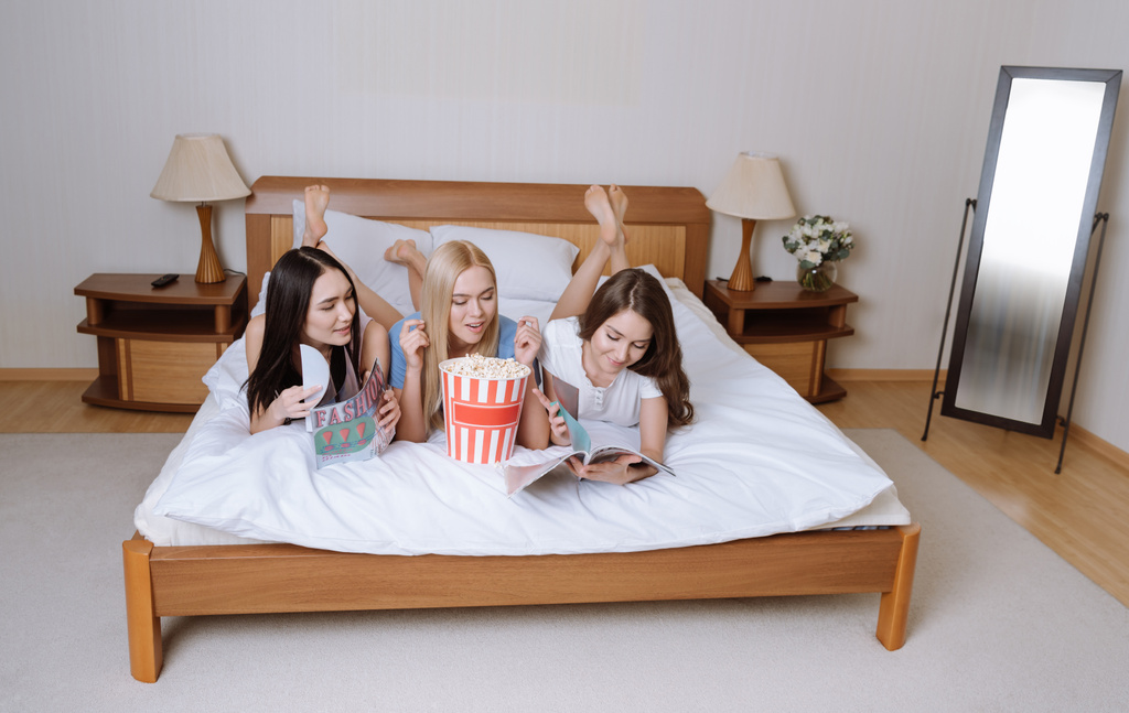 happy multiethnic girls lying on bed with popcorn and reading magazines - Photo, Image