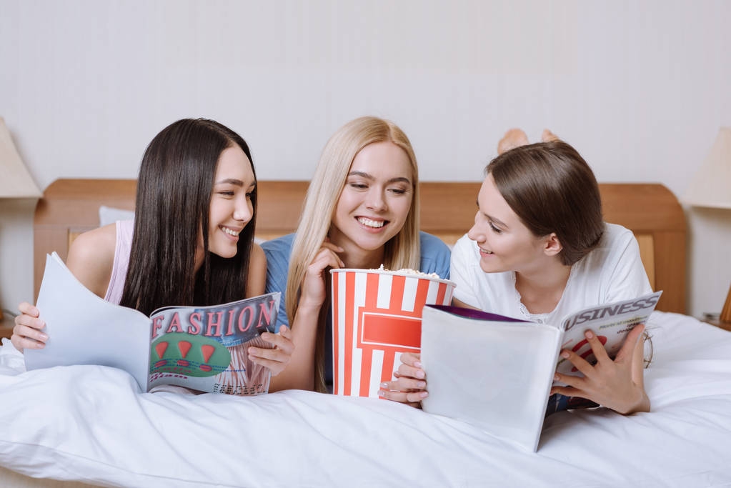 multiethnic friends lying on bed with popcorn and reading magazines - Photo, Image