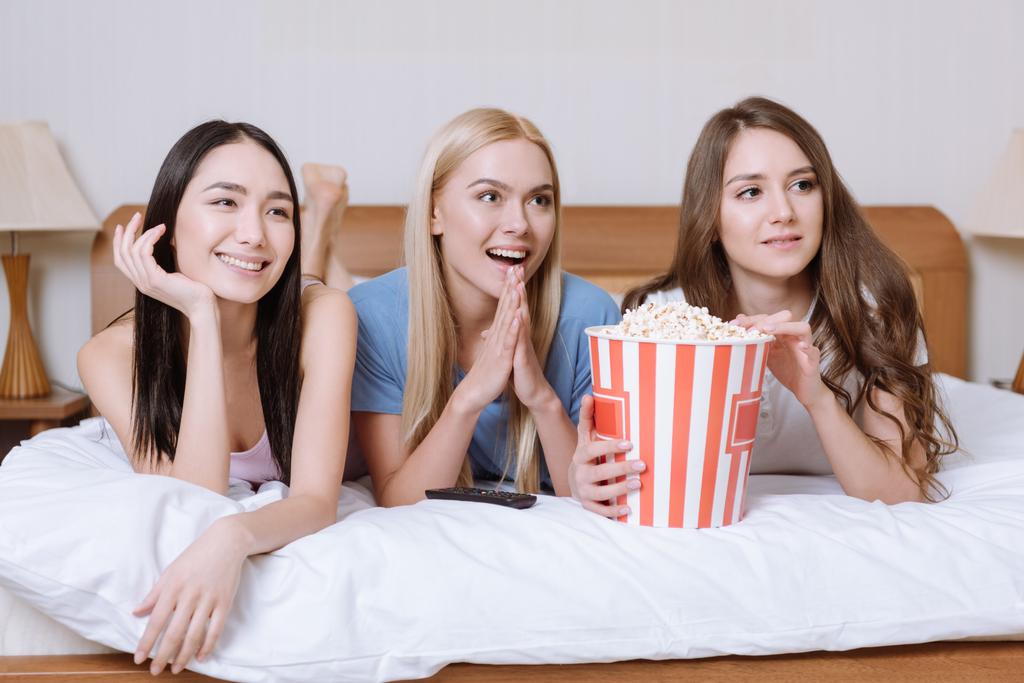 chicas multiétnicas tumbadas en la cama con palomitas de maíz y viendo la televisión
 - Foto, imagen