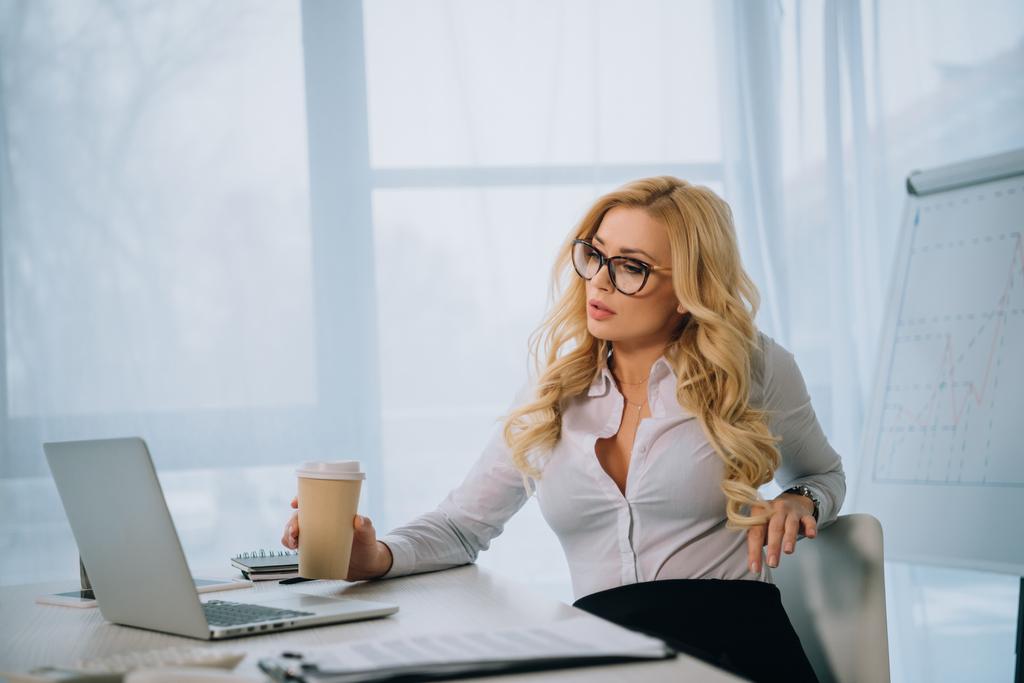 seductive businesswoman holding coffee in paper cup and looking at laptop - Photo, Image