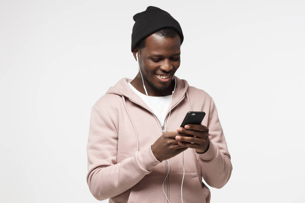 Jonge knappe zwarte man in roze hoodie en zwarte hoed, luisteren van muziek met een koptelefoon en kijken naar scherm van zijn smartphone - Foto, afbeelding