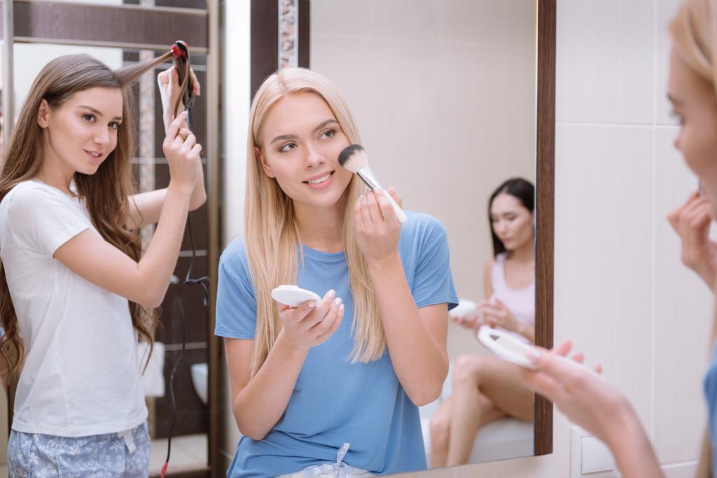 приваблива дівчина застосовує порошок фундаменту з пензлем для макіяжу у ванній кімнаті з друзями
 - Фото, зображення