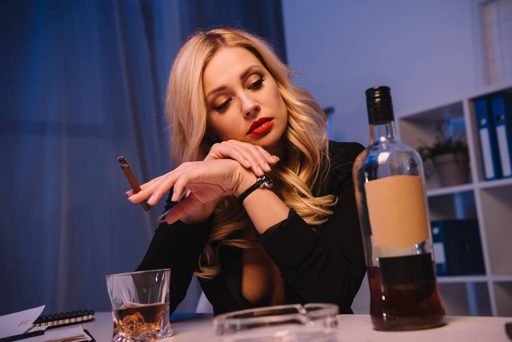 соблазнительная женщина сидит с сигарой и смотрит на бутылку виски в офисе
 - Фото, изображение