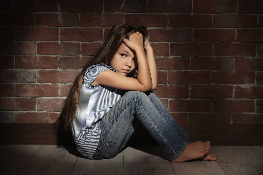 Маленькая грустная девочка, сидящая на полу напротив кирпичной стены. Злоупотребление детской концепцией
 - Фото, изображение