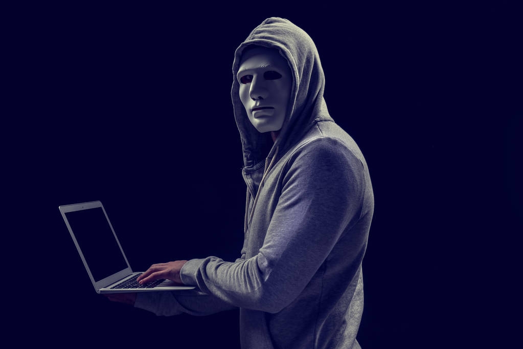 Hacker in masker met laptop op donkere achtergrond - Foto, afbeelding