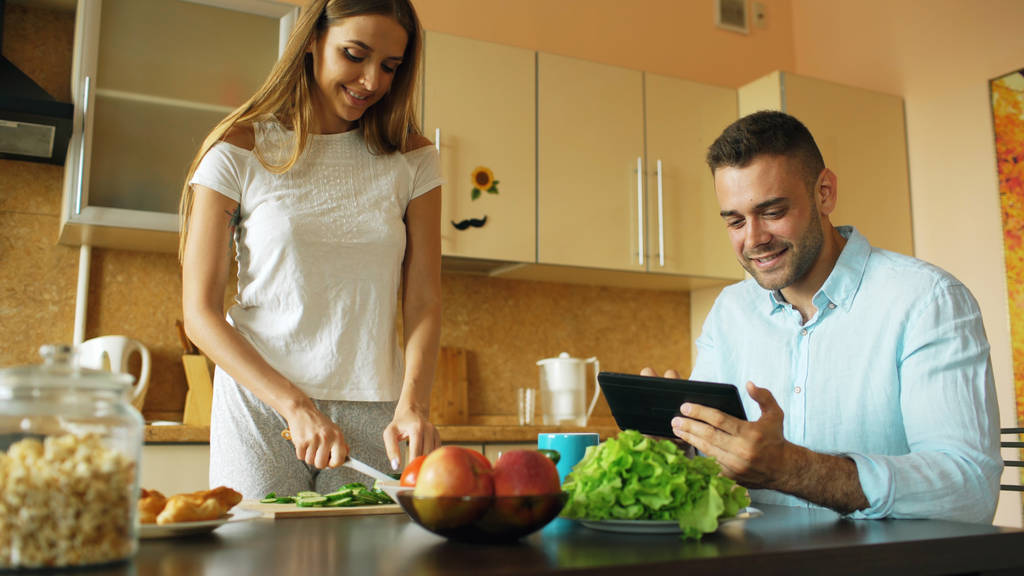 Una coppia attraente che chiacchiera in cucina la mattina presto. Bello uomo utilizzando tablet mentre la sua ragazza cucina
 - Foto, immagini