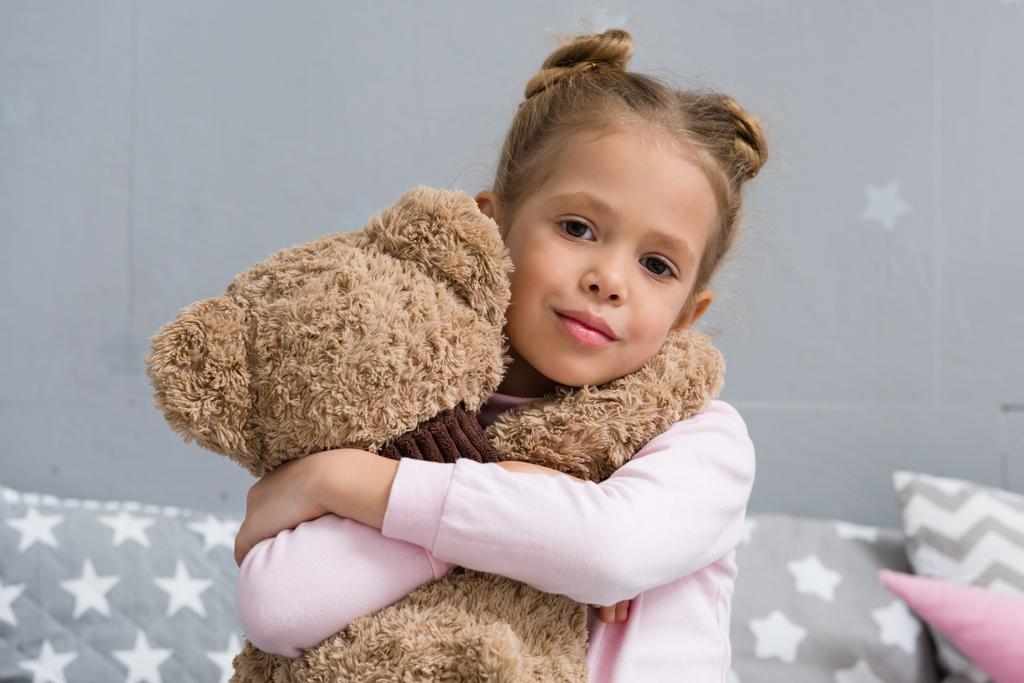 entzückendes kleines Kind umarmt ihren Teddybär und blickt in die Kamera - Foto, Bild
