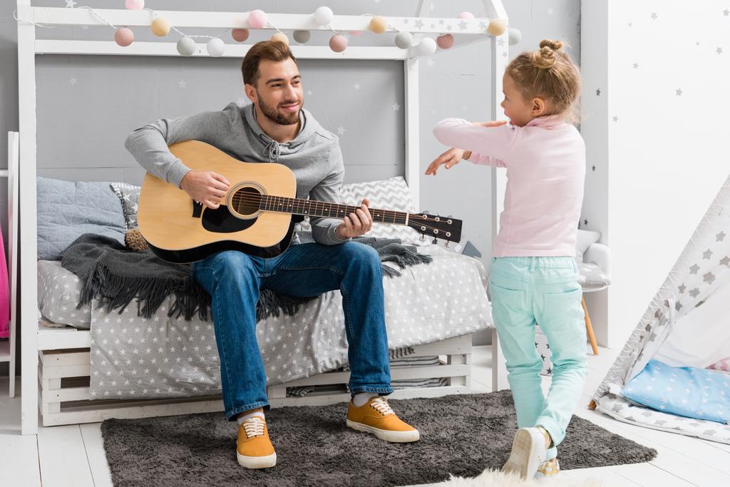 glücklicher Vater spielt Gitarre für Tochter im Schlafzimmer, während sie vor ihm tanzt - Foto, Bild