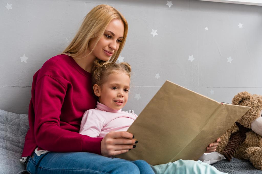 young mother and daughter sitting on bed and reading book together - Photo, Image