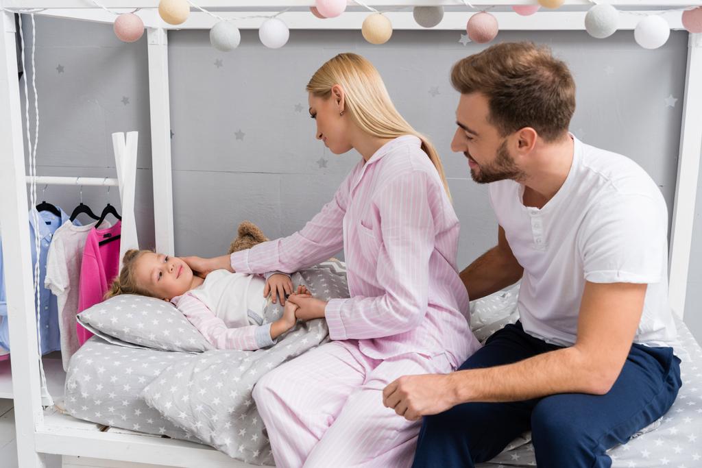 счастливые молодые родители разговаривают с дочерью, пока она лежит в постели в детской спальне
 - Фото, изображение