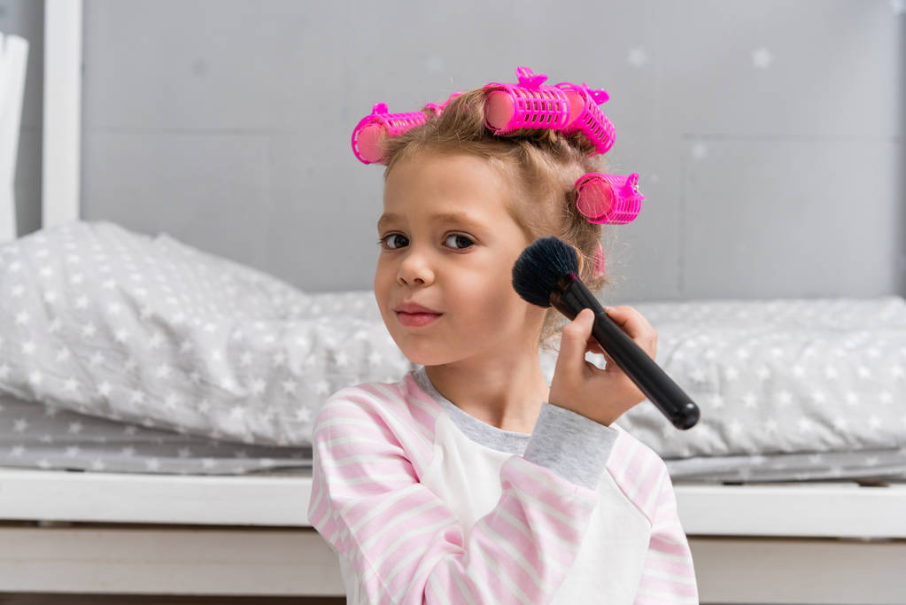 entzückendes kleines Kind mit Haarrollen auf dem Kopf beim Make-up mit Pinsel - Foto, Bild