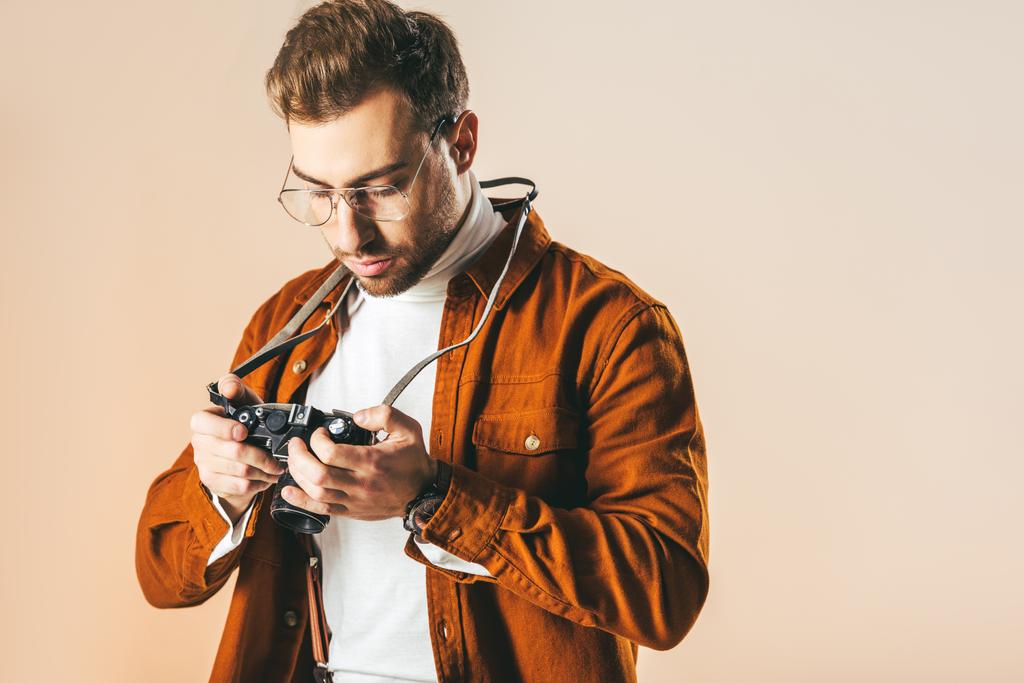 портрет сфокусированного стильного мужчины, смотрящего на фотокамеру в руках, изолированных на бежевом
 - Фото, изображение