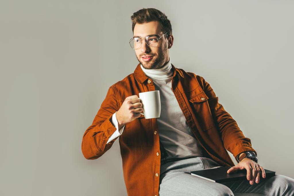 портрет стильного мужчины с чашкой кофе в руке, смотрящего вдаль, изолированного на сером
 - Фото, изображение