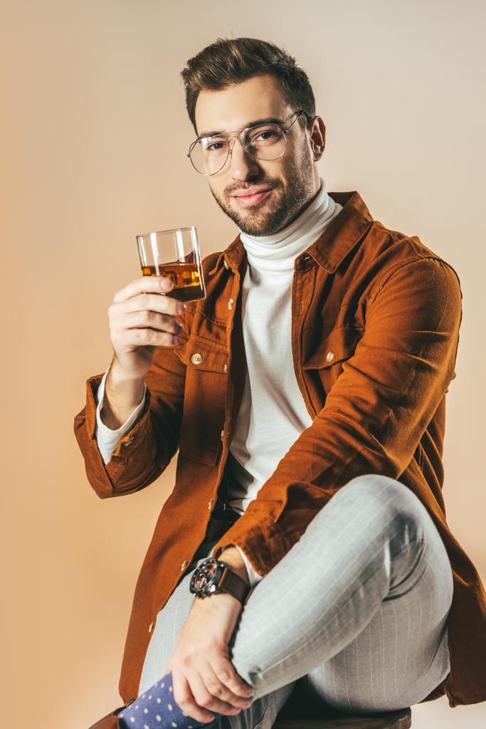 Porträt eines lächelnden, stilvollen Mannes mit einem Glas Cognac isoliert auf beige - Foto, Bild