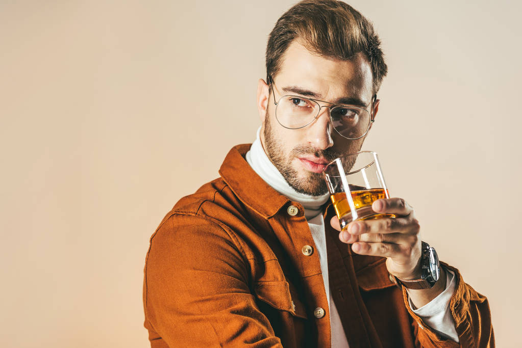 retrato de hombre de moda pensativo con vaso de whisky mirando hacia otro lado aislado en beige
 - Foto, Imagen