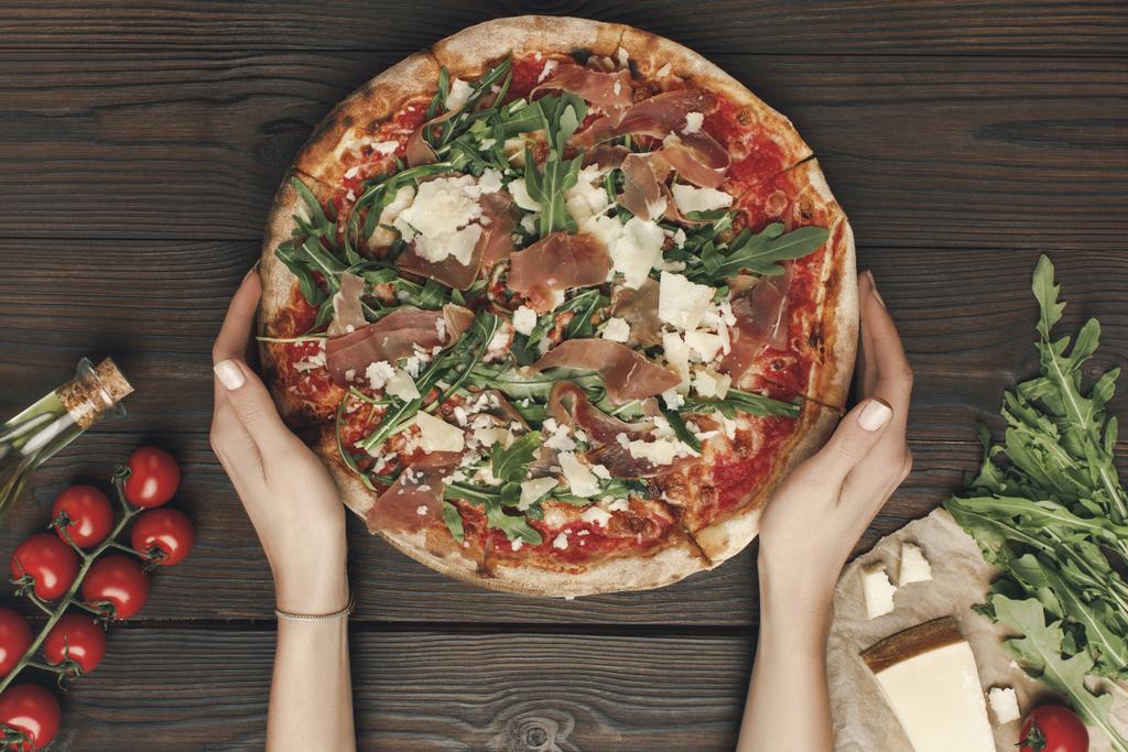 plan recadré d'une femme tenant une pizza maison avec des ingrédients sur une table en bois
 - Photo, image