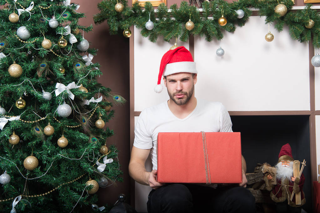 Macho mit Weihnachtsmütze hält verpacktes Paket am Kamin - Foto, Bild