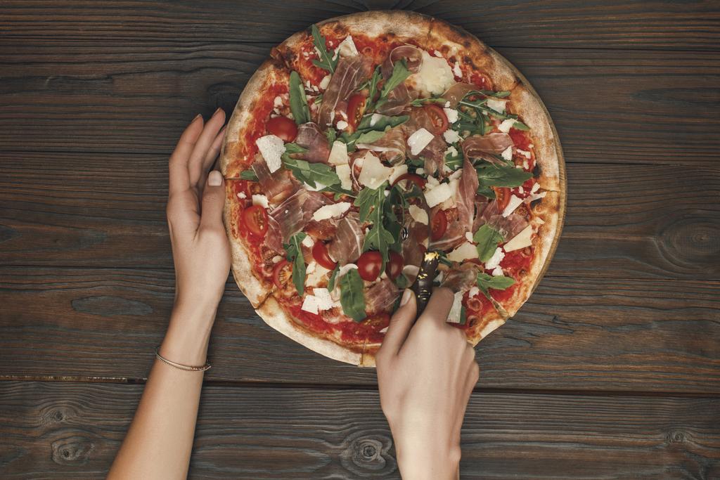 обрезанный снимок женщины, разрезающей итальянскую пиццу на деревянной поверхности
 - Фото, изображение