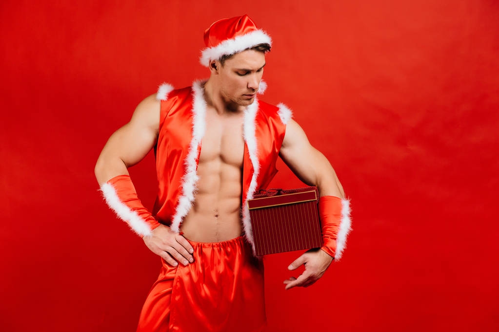 Διακοπές σε γιορτές, Πρωτοχρονιά, Χριστούγεννα. Σέξι γυμναστήριο Αϊ-Βασίλη σε ένα καπέλο κρατά ένα κουτί δώρου. - Φωτογραφία, εικόνα