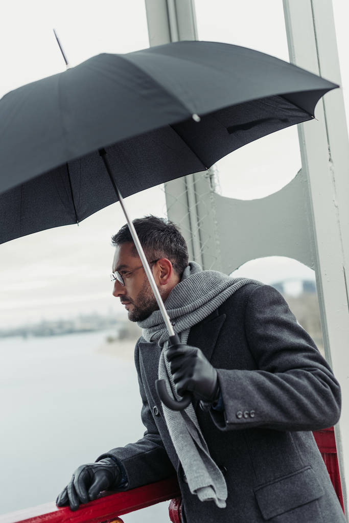 stijlvolle volwassen man in sjaal en jas met paraplu op brug - Foto, afbeelding