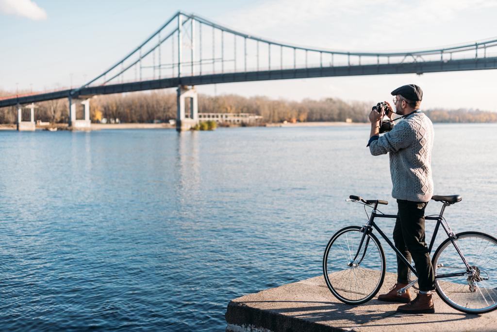 красивий чоловік з старовинною кінокамерою і велосипедом, що стоїть на березі річки
 - Фото, зображення