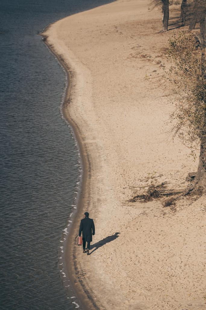pohled z vysokého úhlu člověka v kabátě s kufrem pěšky od písečné pláže na podzim - Fotografie, Obrázek