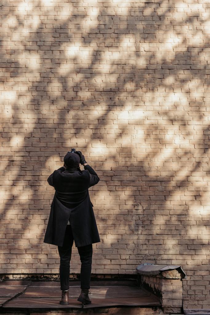 Achteraanzicht van stijlvolle man met camera foto van brrick muur met boom schaduwen - Foto, afbeelding