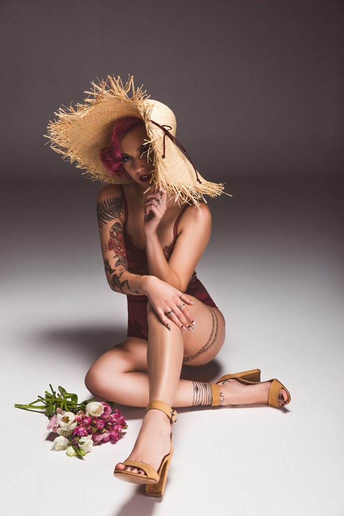beautiful tattoed pin up girl in lingerie and straw hat with flowers infront of grey background  - Photo, Image