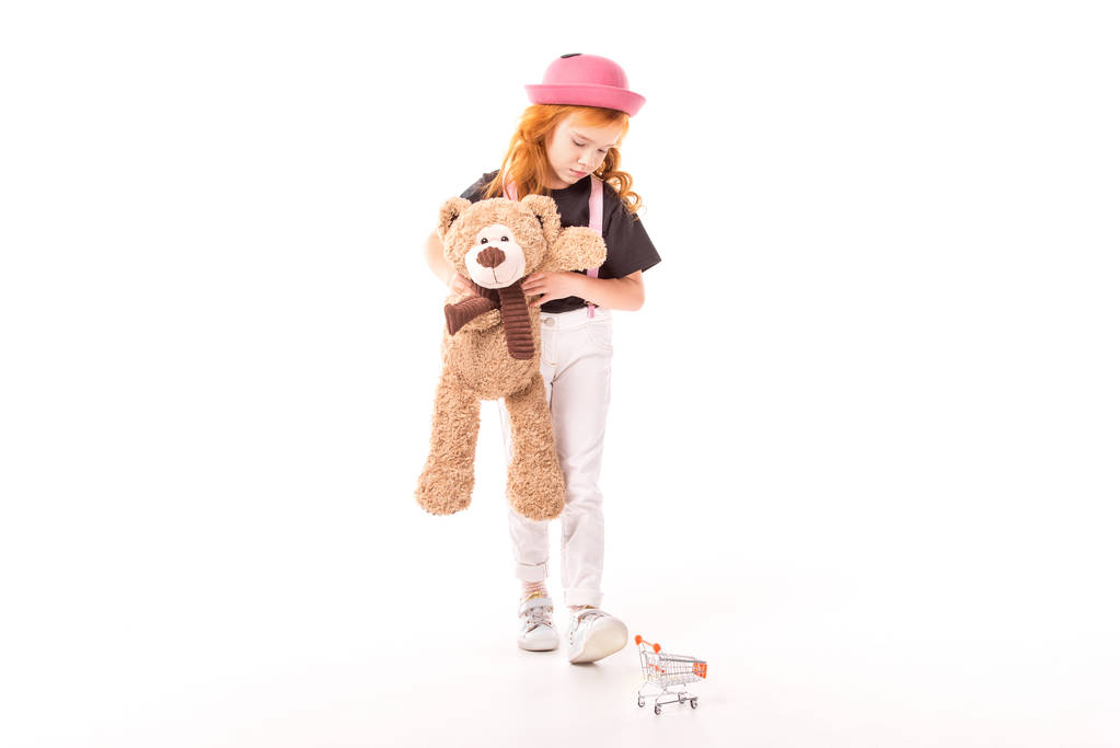 Aufgebrachtes Kind hält Teddybär in der Hand und schaut auf Einkaufswagen-Spielzeug auf weißem Grund - Foto, Bild