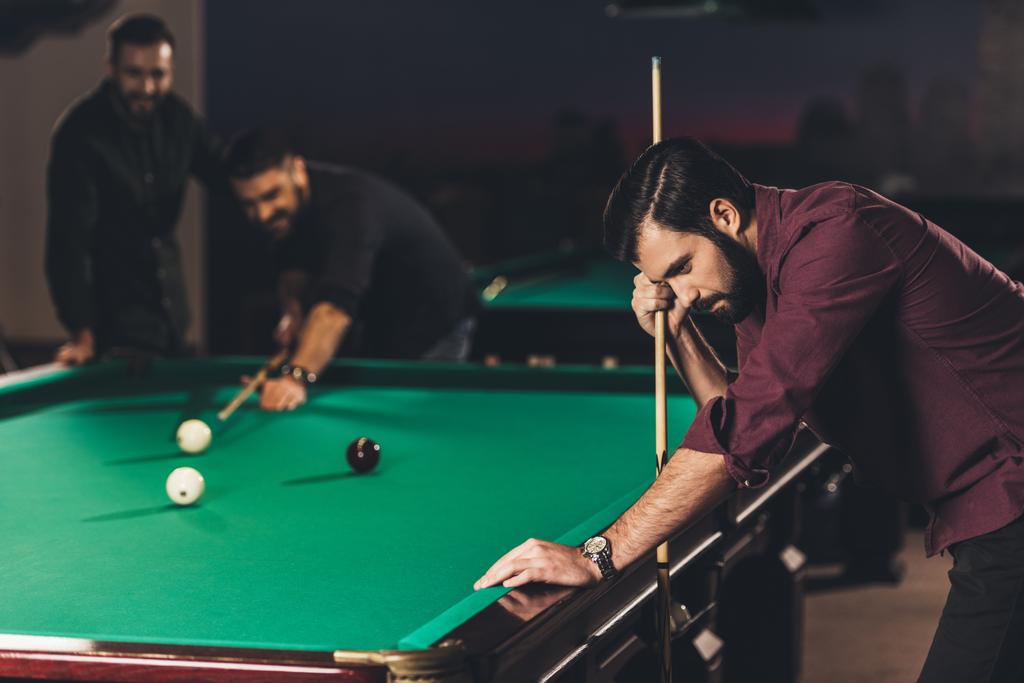 upset caucasian man with cue beside pool table at bar with friends - Photo, Image