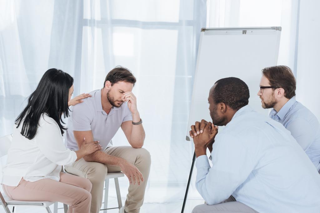 multiethnic middle aged people sitting on chairs and supporting upset man during anonymous group therapy   - Photo, Image