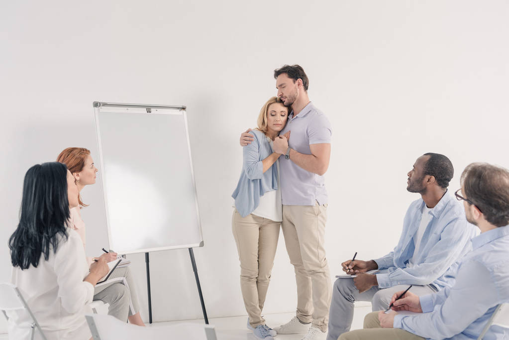 middle aged couple holding hands while standing near blank whiteboard and other people sitting on chairs during group therapy   - Photo, Image