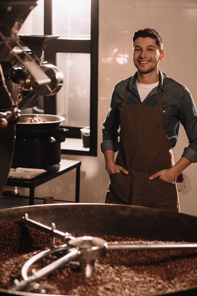 Portret van glimlachen koffie brander staande op roosteren machine en camera te kijken - Foto, afbeelding
