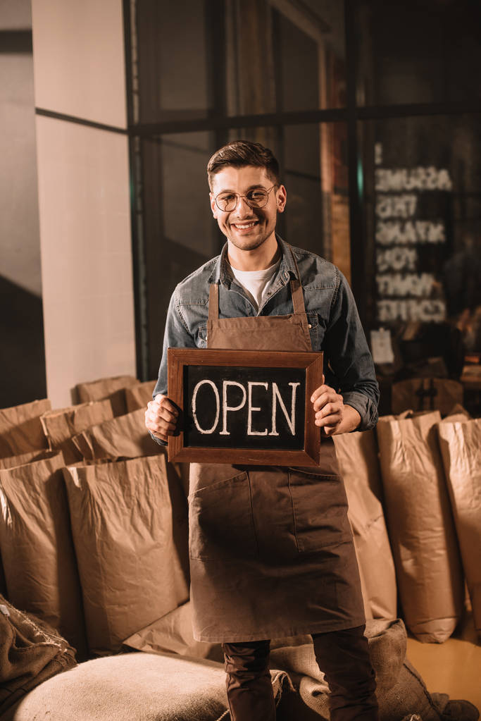 улыбающийся работник кофейни с открытой вывеской, смотрящий в камеру
 - Фото, изображение