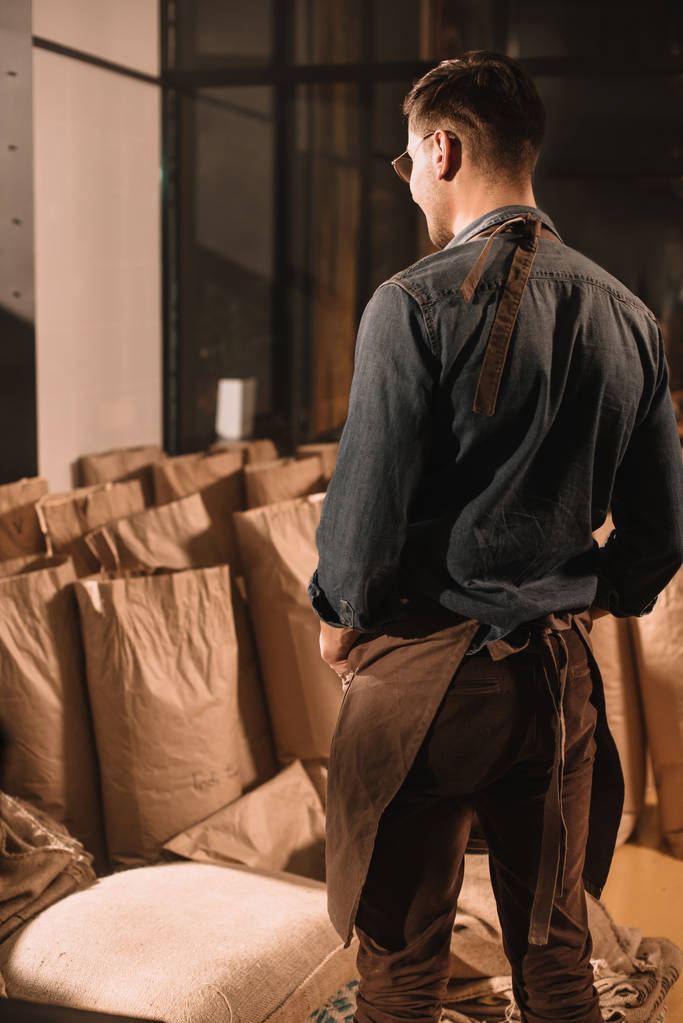 visão traseira do trabalhador da cafetaria no avental olhando para sacos de papel com grãos de café
 - Foto, Imagem