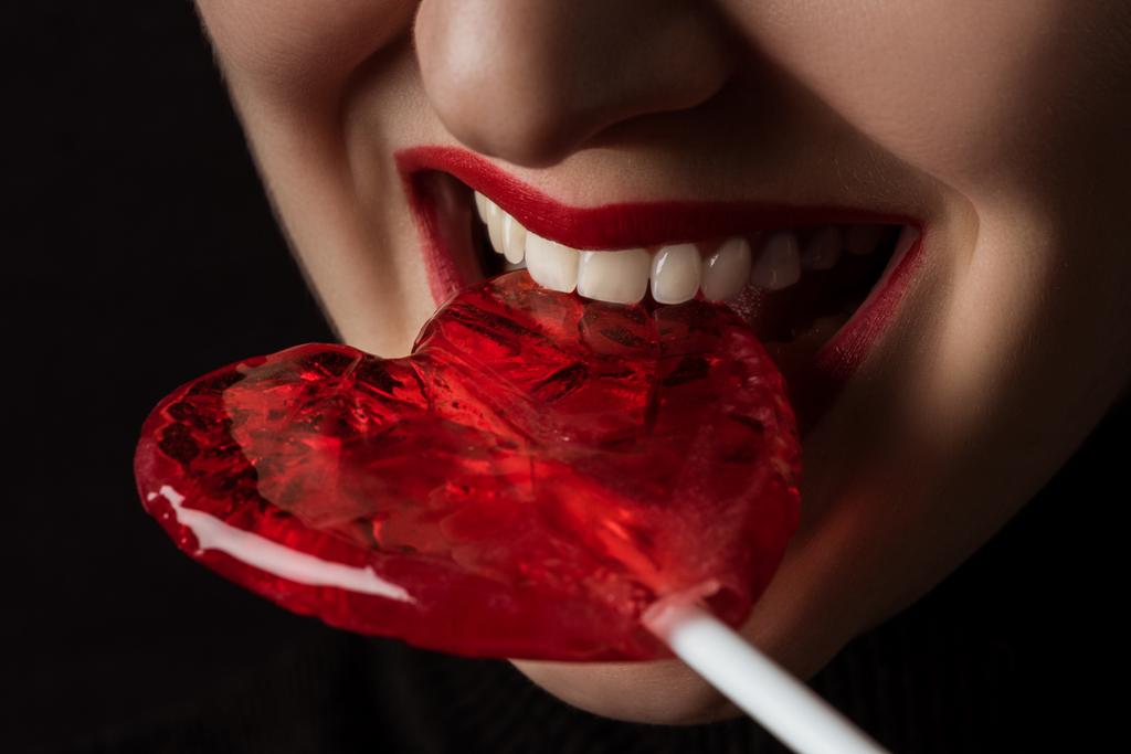 bijgesneden beeld van vrouw bijten hartvormige Lollipop geïsoleerd op zwart, Valentines Day concept  - Foto, afbeelding