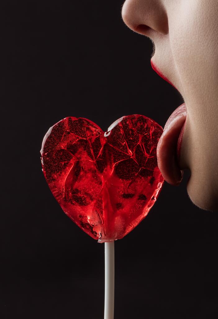 bijgesneden beeld van vrouw likken rood hart gevormde Lollipop geïsoleerd op zwart, Valentines Day concept - Foto, afbeelding