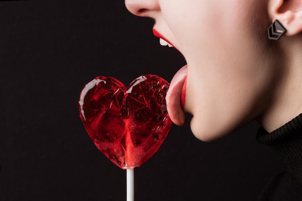 cropped image of woman licking heart shaped lollipop isolated on black, valentines day concept - Photo, Image
