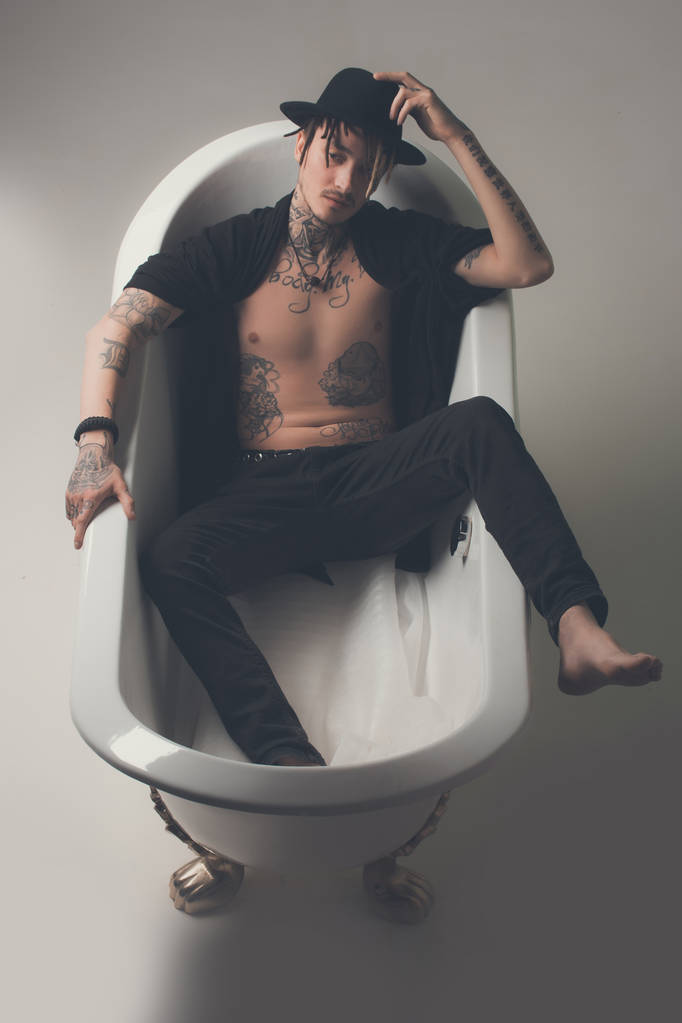 Hombre con sombrero negro y ropa acostado en la pestaña del baño
 - Foto, Imagen